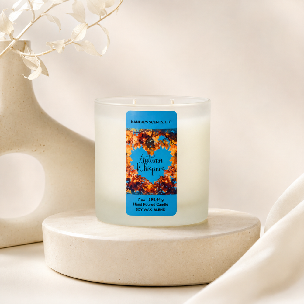 A frosted glass candle jar labeled 'Autumn Whispers' by Kandie's Scents, displayed on a round beige pedestal with a ceramic vase and dried leaves in the background.