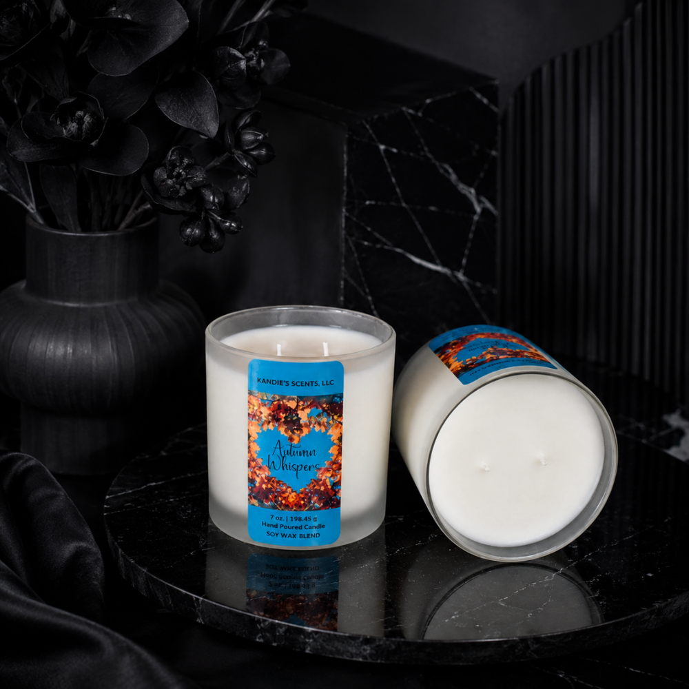 Two white soy candles in a frosted glass jars on a glossy black marble surface, one upright with a blue and orange label reading 'Autumn Whispers' and one lying on its side. A black vase with dark flowers sits in the background.