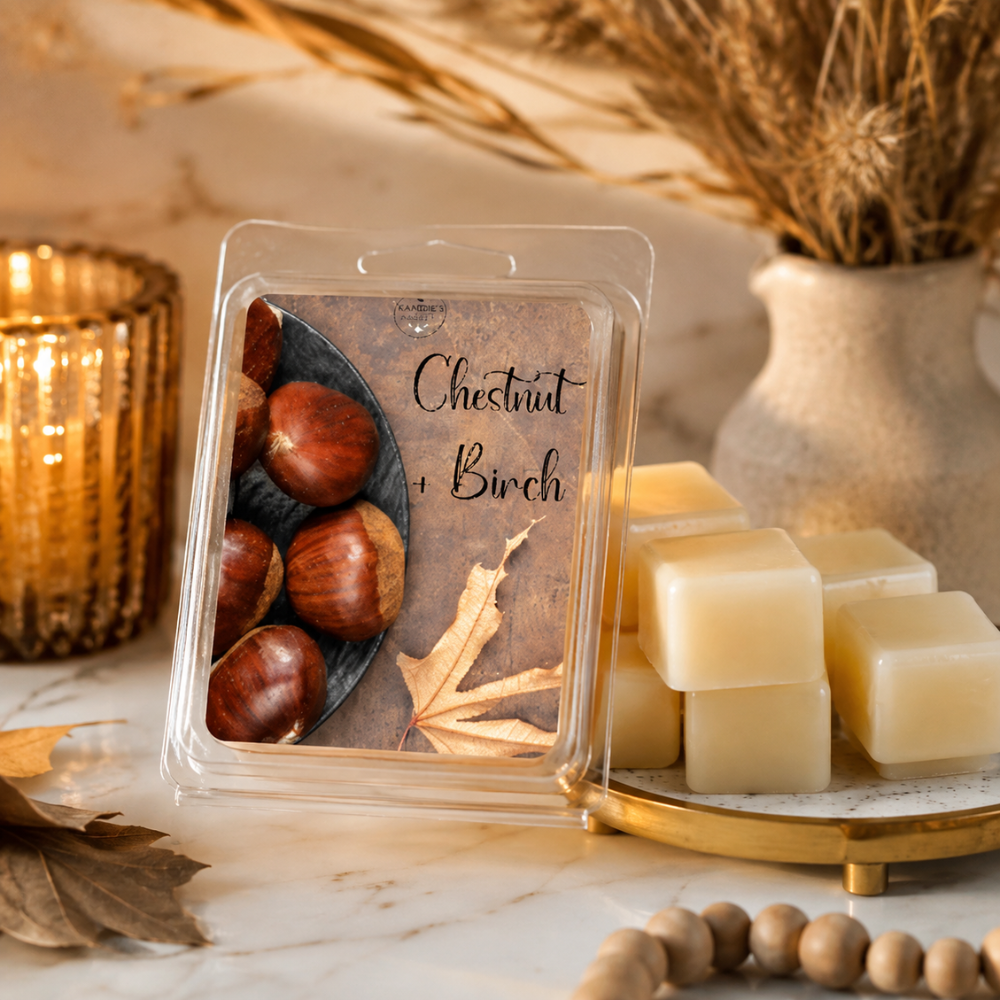 A clear clamshell package labeled 'Chestnut + Birch' sits on a marble surface beside pale cream wax cubes on a gold tray, with autumn decor including dried grass, leaves, and a candle in the background.