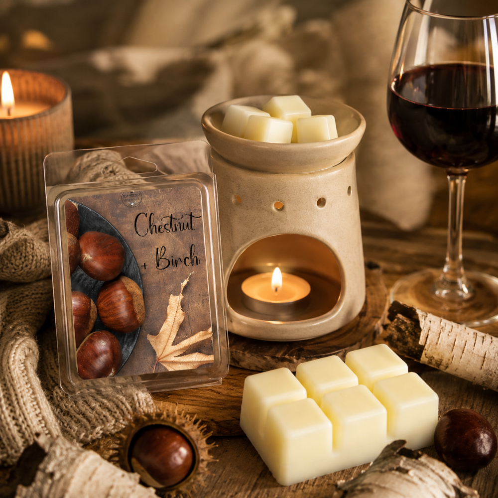A styled product scene showing a clear plastic package labeled 'Chestnut & Birch' wax melts alongside several pale cream wax cubes, a lit candle warmer, a candle, birch bark, and a glass of red wine on a rustic wooden surface.
