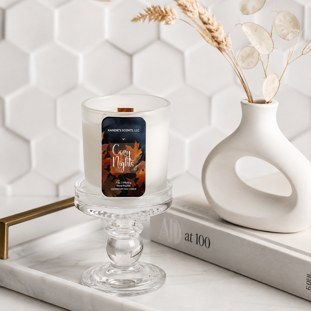 A scented candle in a frosted glass vessel sits on a glass pedestal tray, styled on a marble surface beside a white ceramic vase with dried botanicals and a book. The candle label reads 'Kandie's Scents' and 'Cozy Nights.