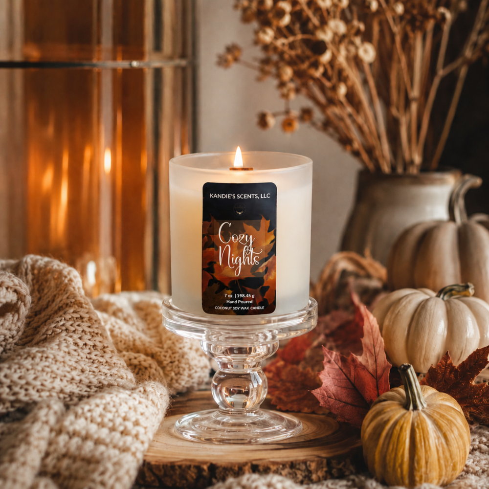 A lit white candle in a frosted glass pedestal holder, labeled 'Cozy Nights' by Kandie Scents. The candle is styled in a warm autumn setting with dried flowers, small pumpkins, red leaves, and a knitted blanket.