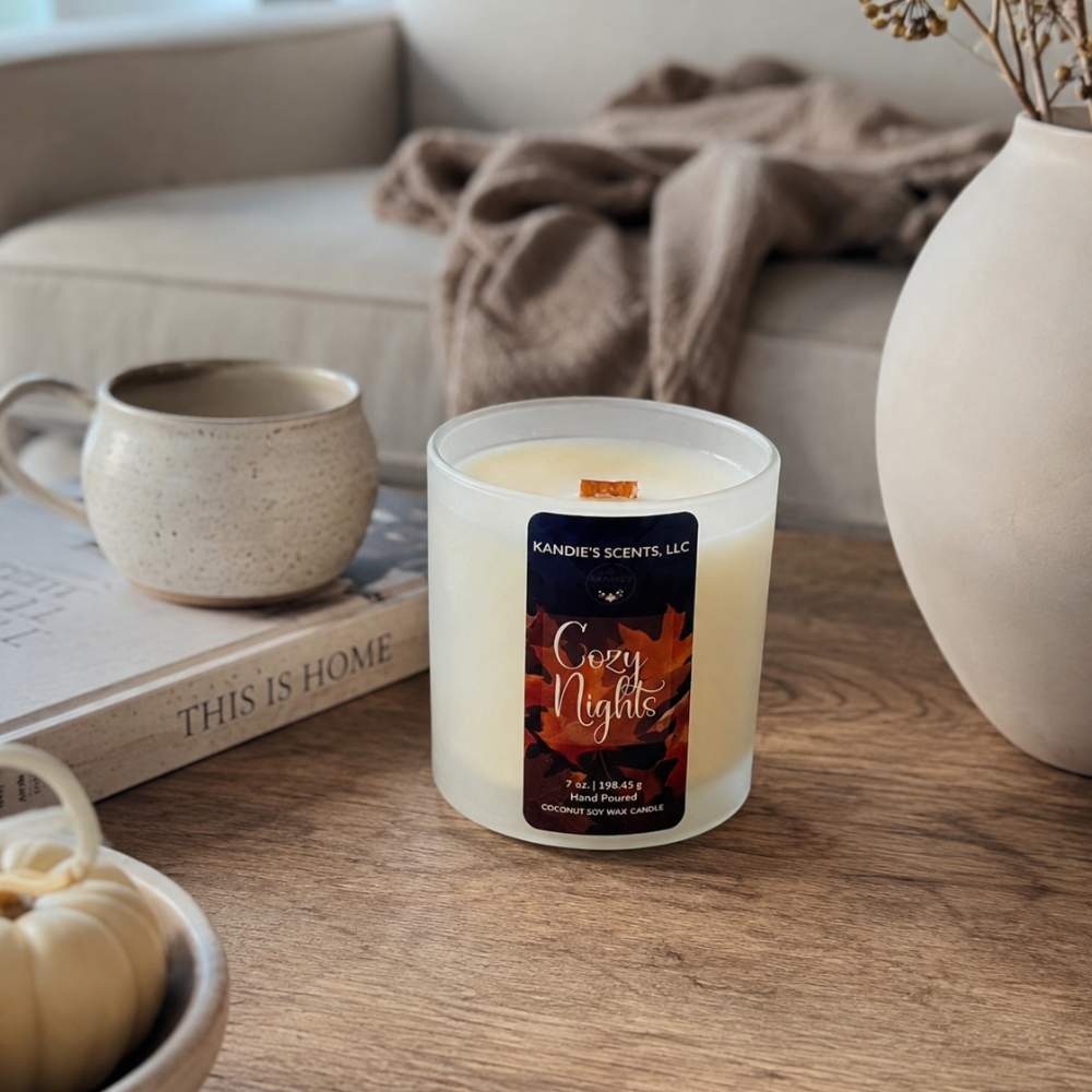 A frosted glass jar candle labeled 'Cozy Nights' by Kandie's Scents, placed on a wooden table in a cozy living room setting with a mug, book, and vase in the background.