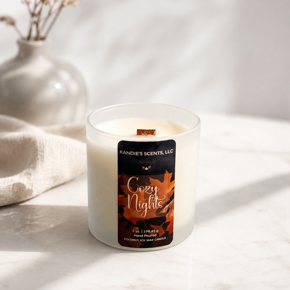 A frosted glass candle jar labeled 'Kandie's Scents. Cozy Nights' sits on a light marble surface with a soft neutral fabric and vase in the background.