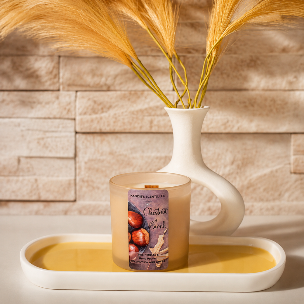 A scented candle in a tan glass jar sits on a white tray in front of a white ceramic vase holding dried golden grass, with a beige stone-tile wall background.