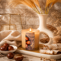 A lit candle in a clear glass jar sits on a tray in a styled autumn scene with dried grass in a white vase, a bowl of chestnuts, birch bark, and a beige linen cloth. The candle label reads 'Kandie's Scents' and 'Chestnut Birch'.