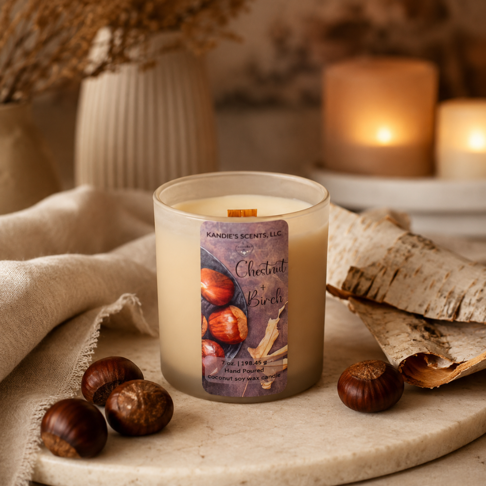 A candle in a tan glass jar sits on a stone tray surrounded by chestnuts, birch bark, and a beige cloth, with warm blurred candles in the background. The label reads 'Kandie's Scents' and 'Chestnut + Birch'.