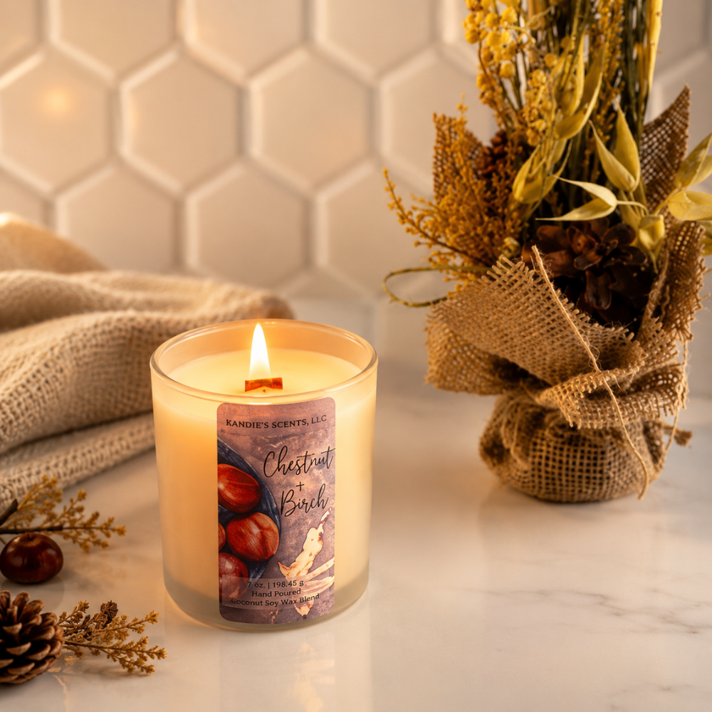 A lit candle in a clear glass jar sits on a marble surface, surrounded by dried flowers in burlap, a beige cloth, and pinecones. The candle label reads 'Chestnut + Birch' and shows the brand KANDIE'S SCENTS.