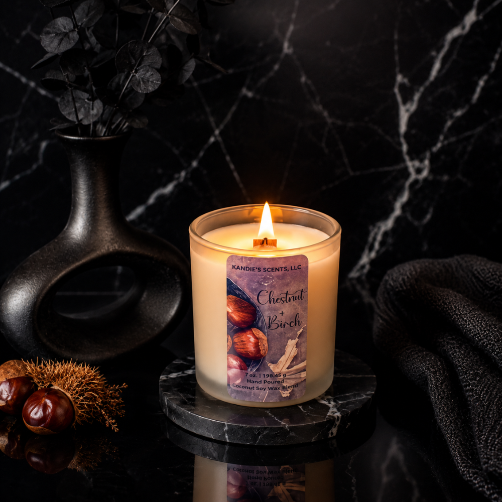 A lit candle in a tan glass jar sits on a small round marble coaster against a dark marble background. The label reads 'Kandie's Scents' and 'Chestnut + Birch.