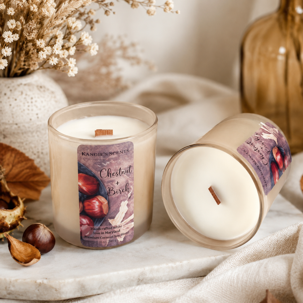 Two candles in glass jars on a marble surface with dried flowers, leaves, and a bottle in a styled setting. One candle is upright with a visible front label reading 'Kandie's Scents Chestnut + Birch,' and the other is tipped on its side showing the wax top and a small wooden wick.