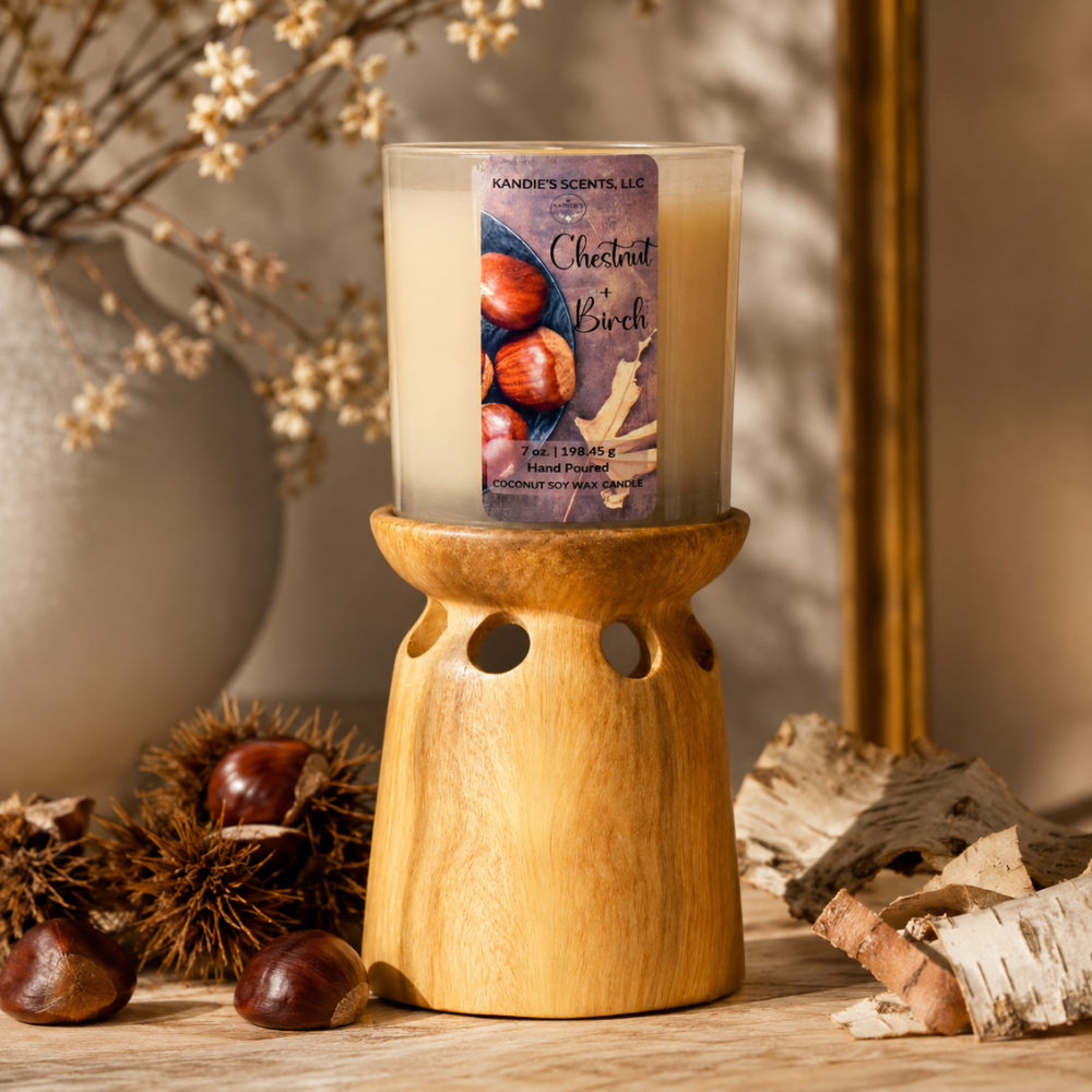 A scented candle in a tan glass jar sits on a wooden holder. The candle label reads 'KANDIE'S SCENTS' and 'Chestnut + Birch.