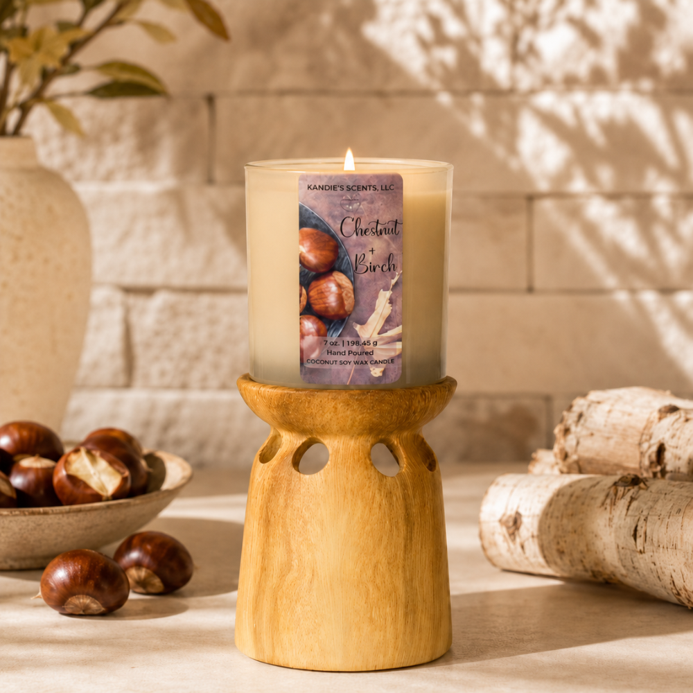 A lit coconut candle in a clear glass jar sits on a wooden scented candle holder. The candle label reads 'KANDIE'S SCENTS.