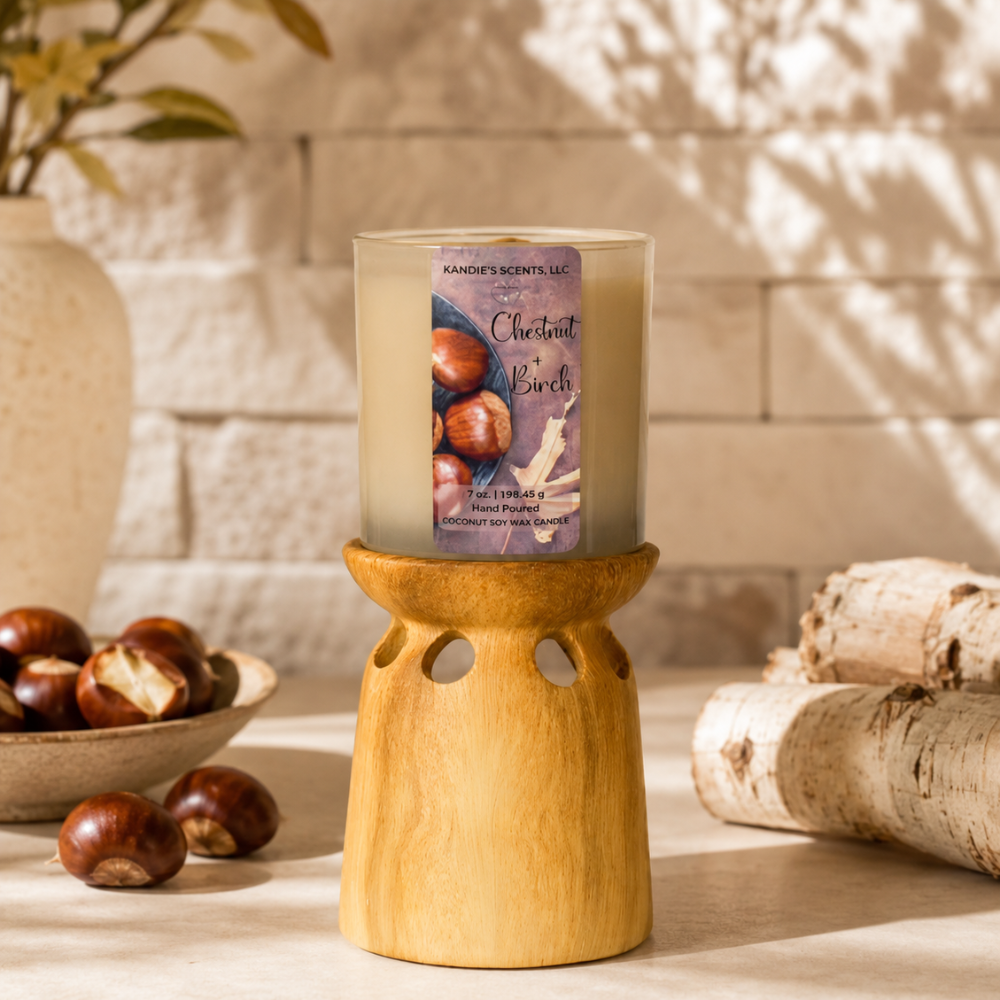 A scented candle in a tan glass jar sits on a light natural wood holder. The candle label reads 'Chestnut + Birch' and shows hazelnuts and wood.
