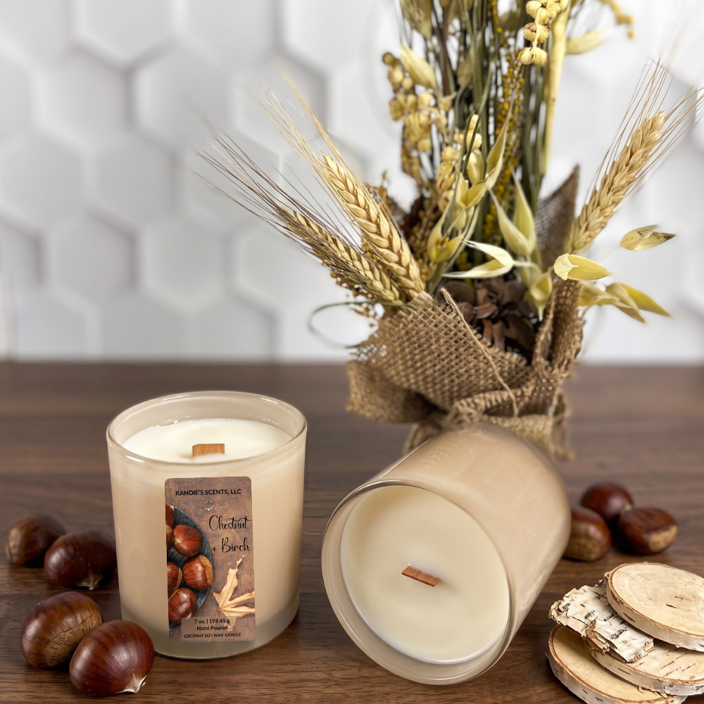 A scented candle shown in two views on a wooden surface: one upright with a tan glass jar and cream wax, and one tipped on its side showing the beige vessel. The label reads Chestnut + Birch.