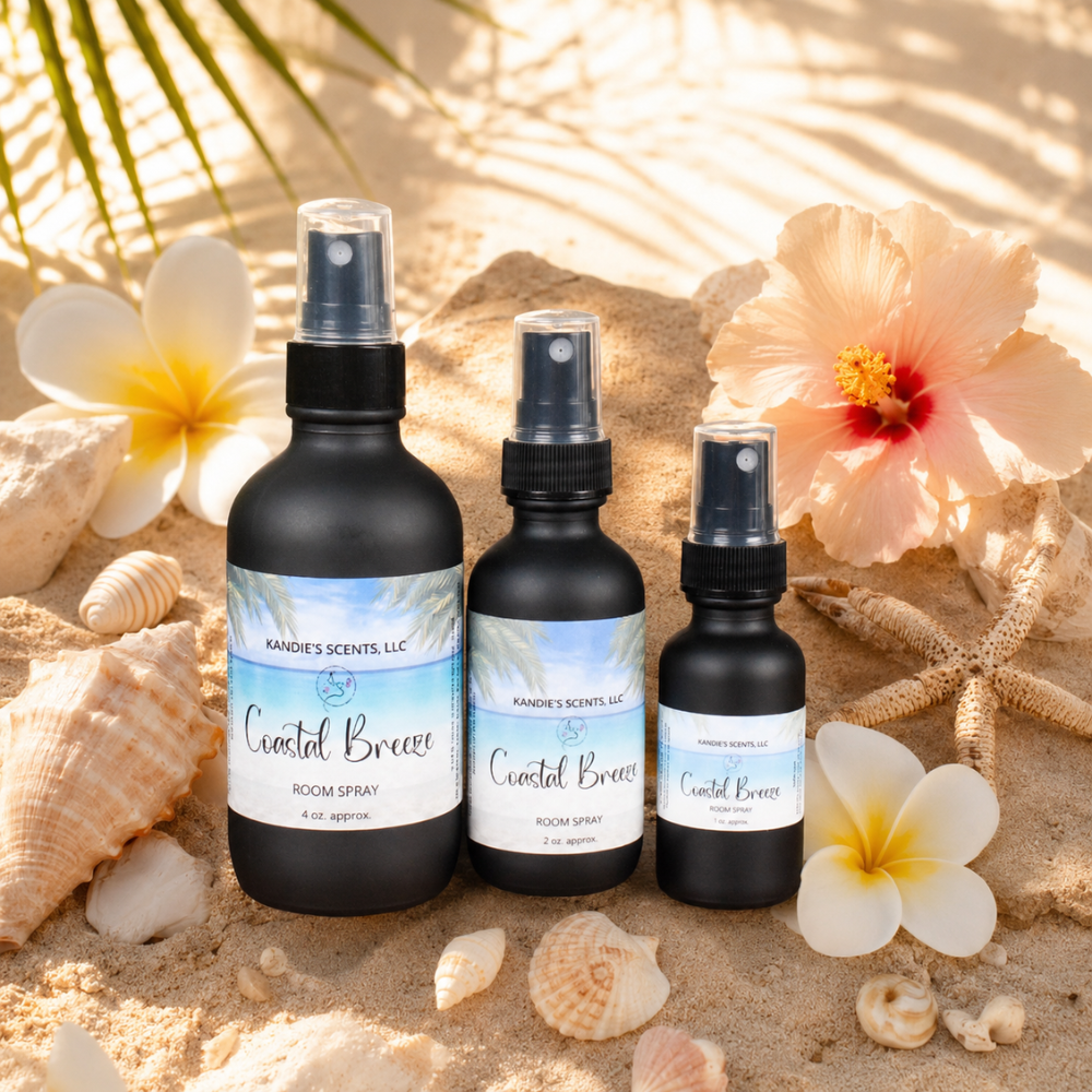 Three 'Coastal Breeze' room spray bottles on sand with flowers and shells.