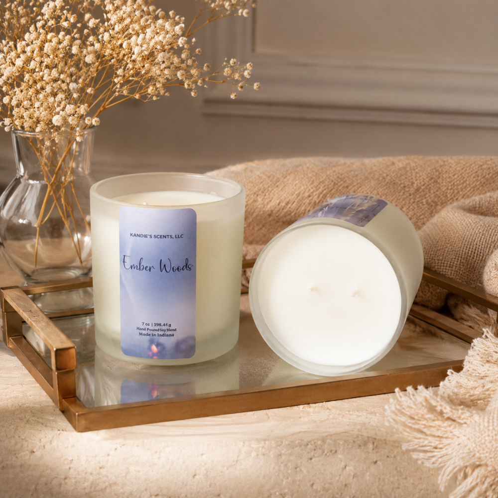 A scented candle in a frosted glass jar sits on a mirrored tray beside its lid, styled with dried flowers and a beige textile in a warm indoor setting. The label reads 'Kandie's Scents' and 'Ember Woods'.