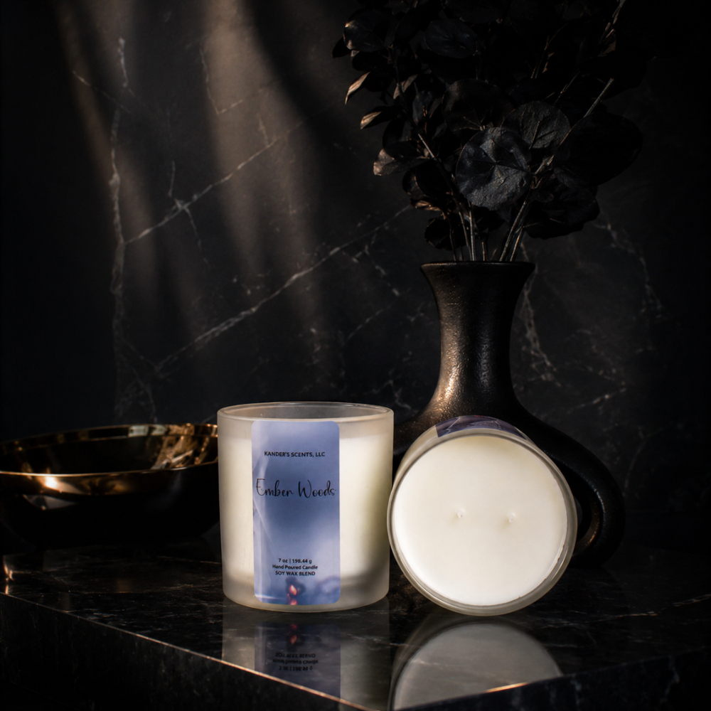 A frosted glass candle jar labeled 'Ember Woods' stands beside its open lid on a dark marble surface, with a black vase of dried dark foliage and a gold bowl in the background.