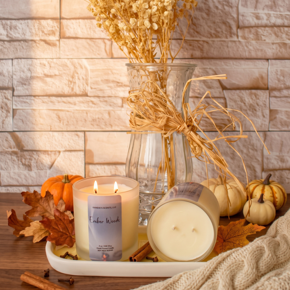 One lit candle and one unlit candle in glass jars displayed on a tray with autumn decor, including pumpkins, leaves, a glass vase with dried flowers, and a knitted blanket, against a light stone wall.