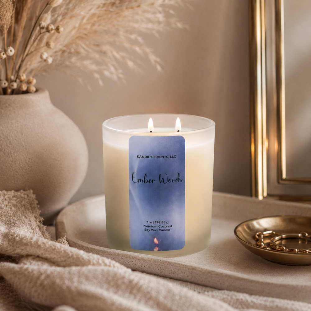 A lit double-wick soy wax candle in a frosted glass jar sits on a beige stone tray in a styled interior setting. The label reads 'Kandie's Scents' and 'Ember Woods'.