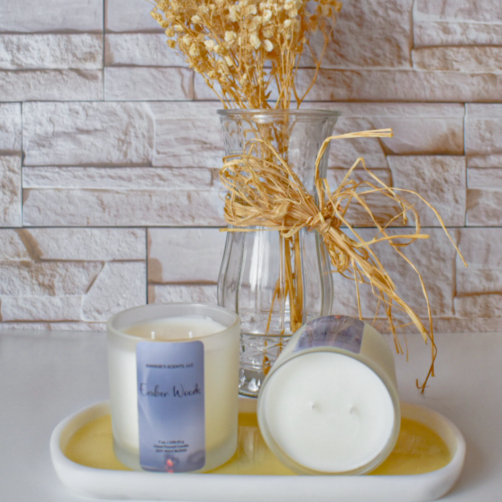 A scented candle in a frosted glass jar with a lavender label, shown alongside another candle lying on its side to display the white wax surface. The candles are arranged on a white tray with a yellow wax drip, set against a glass vase of dried grasses in front of a light stone wall.