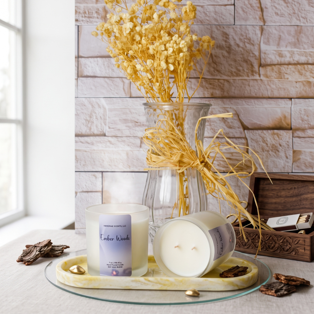 A candle product styled on a glass tray with dried flowers in a vase, set against a stone wall and window. The candle labels read 'Ember Woods'.