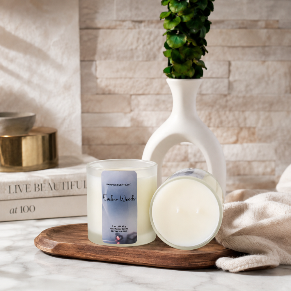A candle set on a wooden tray in a styled home setting. One candle is upright with a clear glass jar and a blue-gray label reading 'Kandie's Scents" and 'Ember Woods'; the other is tipped over to show the creamy off-white wax.