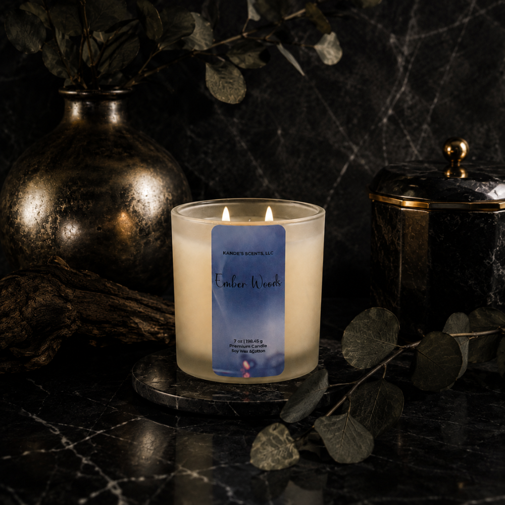 A lit double-wick candle in a frosted glass jar sits on a dark marble surface with decorative eucalyptus leaves, a metallic vase, and a dark lidded container in the background. The front label reads 'Kandie's Scents' and 'Ember Woods'.