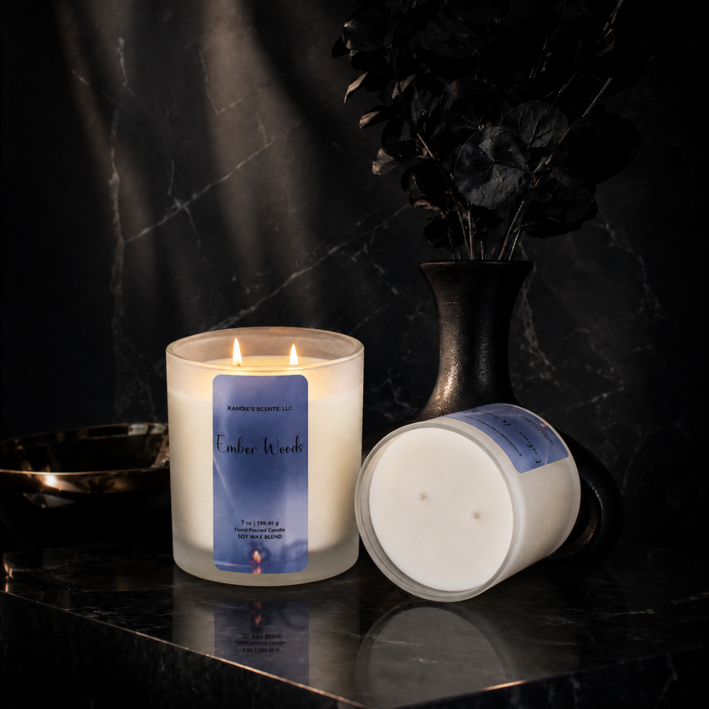 A lit scented candle in a frosted glass jar stands beside its matching lid on a dark marble surface. The label reads 'Ember Woods' and 'Kandie's Scents'.