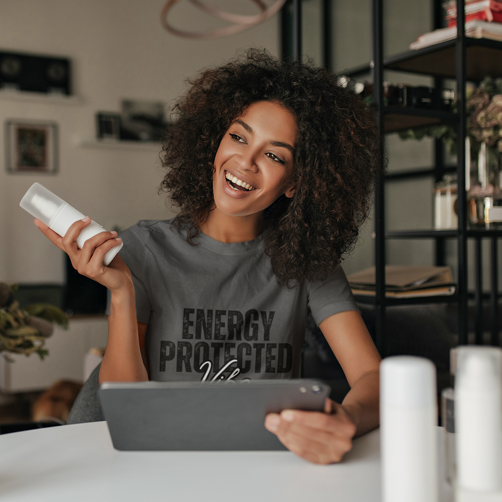Woman sitting at a desk holding a phone and beauty product, wearing a 'Energy Protected' shirt.