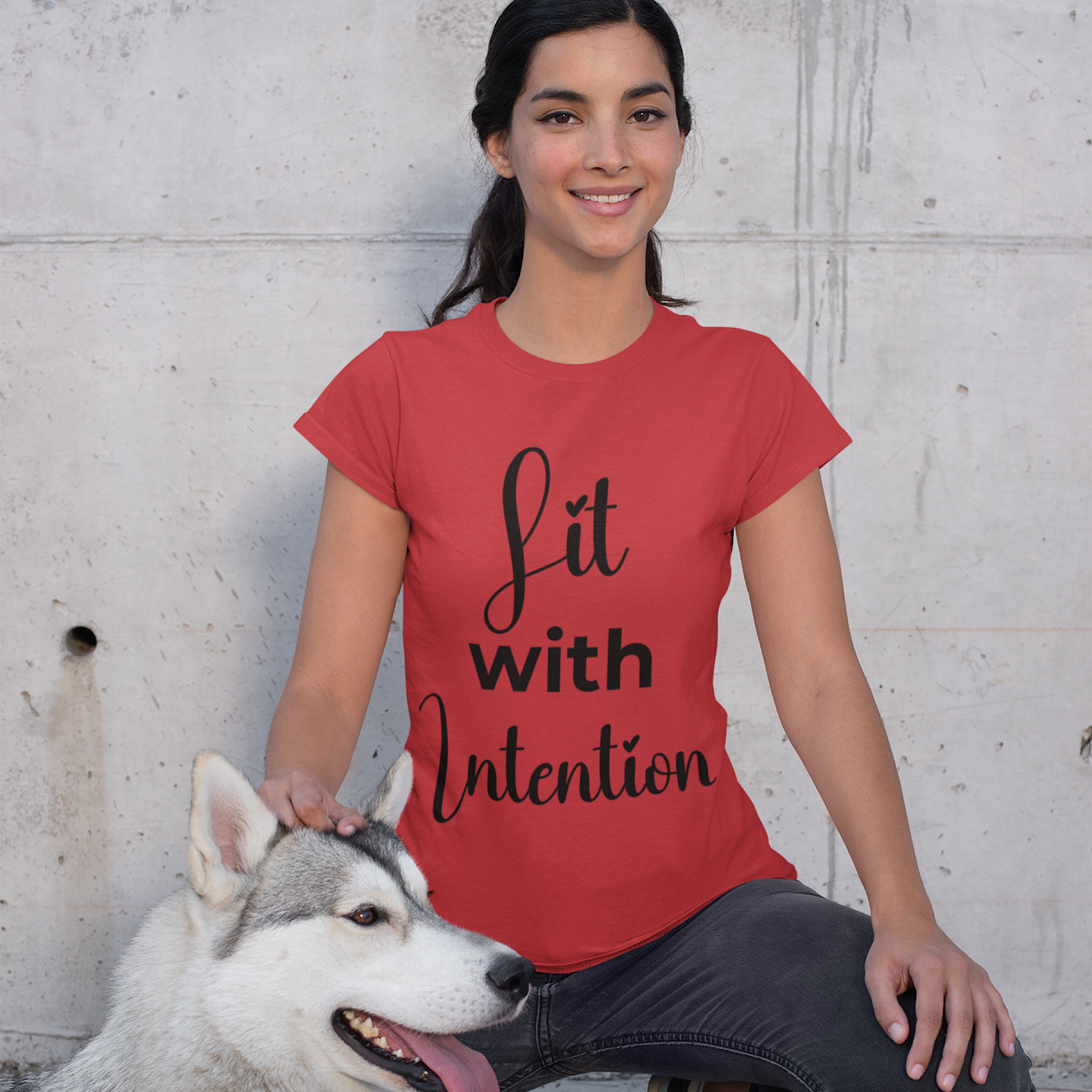 Woman wearing a red t-shirt with 'Lit with Intention' text, sitting next to a dog against a concrete wall.