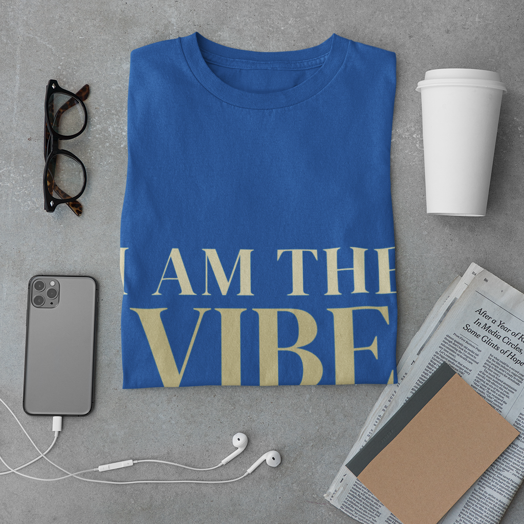 Blue t-shirt with 'I AM THE VIBE' text on a gray surface with phone, coffee, and glasses.