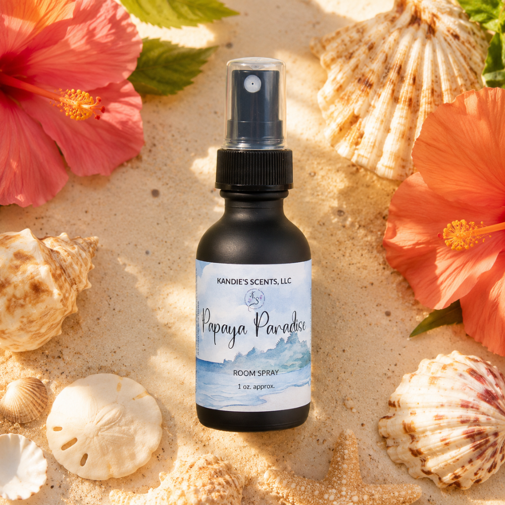 Room spray bottle labeled 'Papaya Paradise' surrounded by seashells and pink flowers on sand.