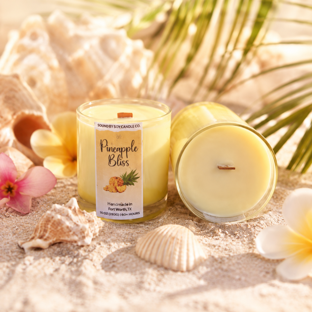 Two candles labeled 'Pineapple Bliss' on sand with shells and flowers