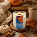 A lit scented candle in a white glass jar sits on a wooden coaster in a cozy autumn setting with a knit blanket, small pumpkins, dried leaves, cinnamon sticks, and a ceramic mug.
