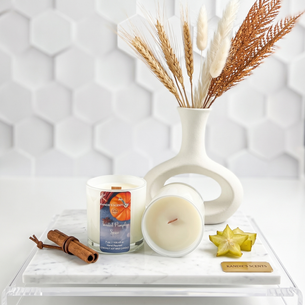 A styled product display featuring a candle in a white glass jar with a label reading 'Toasted Pumpkin Spice' on a marble tray. A small cinnamon stick, starfruit, with a white ceramic vase holding dried wheat and pampas grass behind the candle.