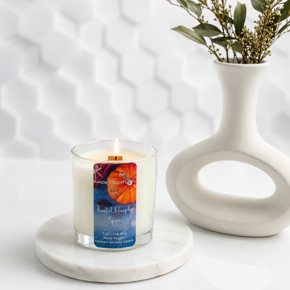 A lit candle in a clear glass jar sits on a round white marble coaster beside a white ceramic vase with dried greenery. The candle label reads 'Kandie's Scents' and 'Toasted Pumpkin Spice.