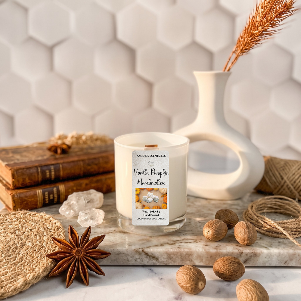 A candle in a white glass jar with a white wax fill and a front label reading 'Vanilla Pumpkin Marshmallow' by Kandie's Scents, styled on a marble board with books, a ceramic vase, twine, crystals, and star anise against a white hexagonal tile background.