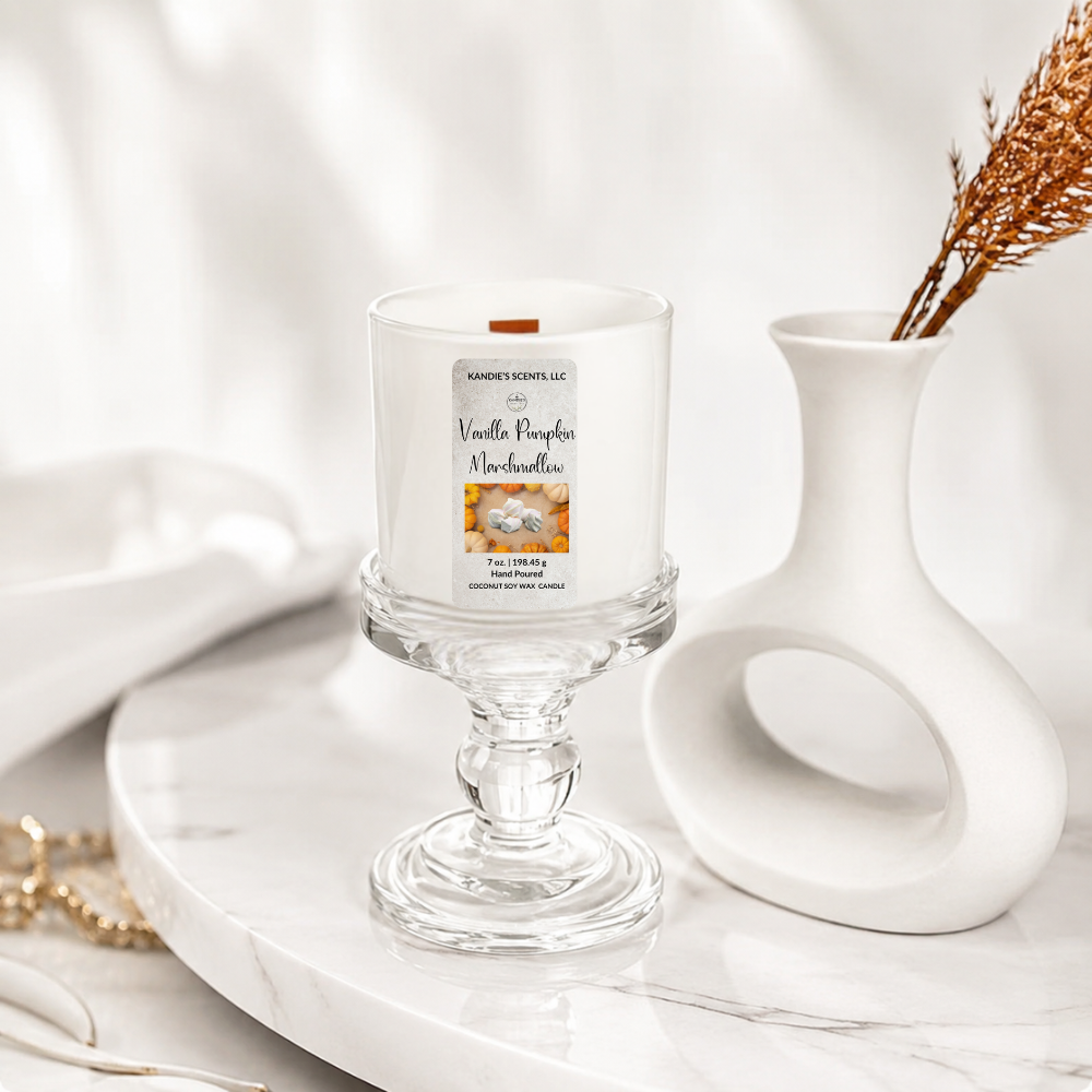 A white scented candle in a white glass pedestal holder on a marble tray, styled beside a white sculptural vase with dried branches.