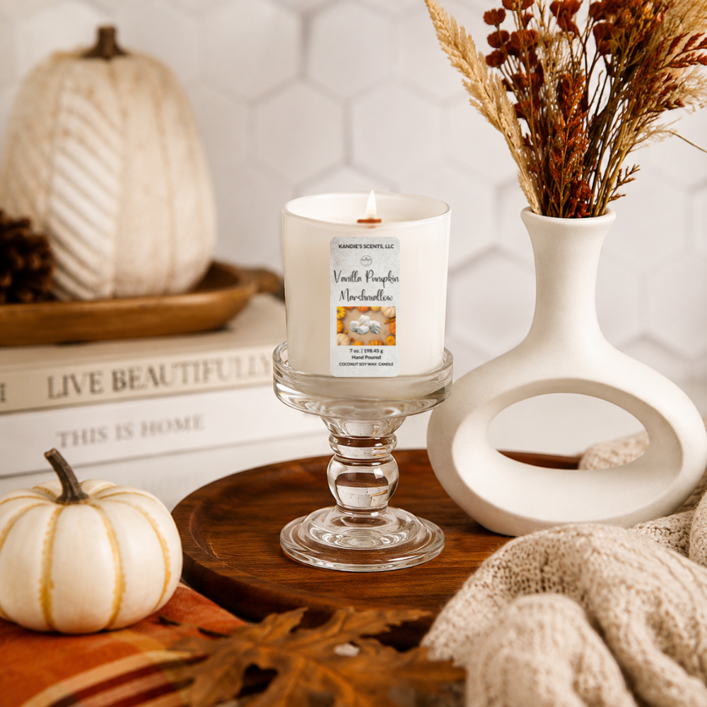A white scented candle in a white glass candlestick holder sits on a round wooden tray, styled in a cozy autumn home setting with a white ceramic vase, dried flowers, a small white pumpkin, and a knitted blanket.