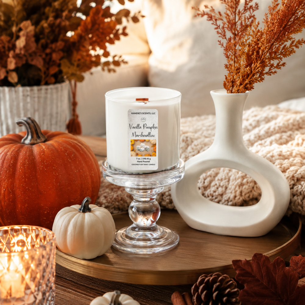 A white scented candle in a white glass pedestal holder, styled on a wooden tray with a small pumpkin, a white vase, dried stems, and a warm candlelit setting.