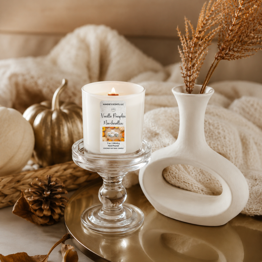 A lit white candle in a white glass pedestal holder, styled on a reflective gold tray beside a white sculptural vase with dried stems, pumpkins, a pinecone, and soft neutral textiles in an autumn-themed setting.