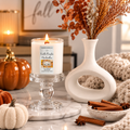 A lit scented candle in a white glass pedestal holder sits on a marble surface in a styled fall setting with pumpkins, dried flowers in a white vase, cinnamon sticks, and a cream knit blanket.