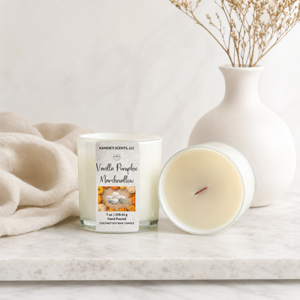A candle in a clear glass jar labeled 'Vanilla Pumpkin Marshmallow' by Kandie's Scents, shown next to its open lid on a marble surface with a cream knit throw and a white vase in the background.