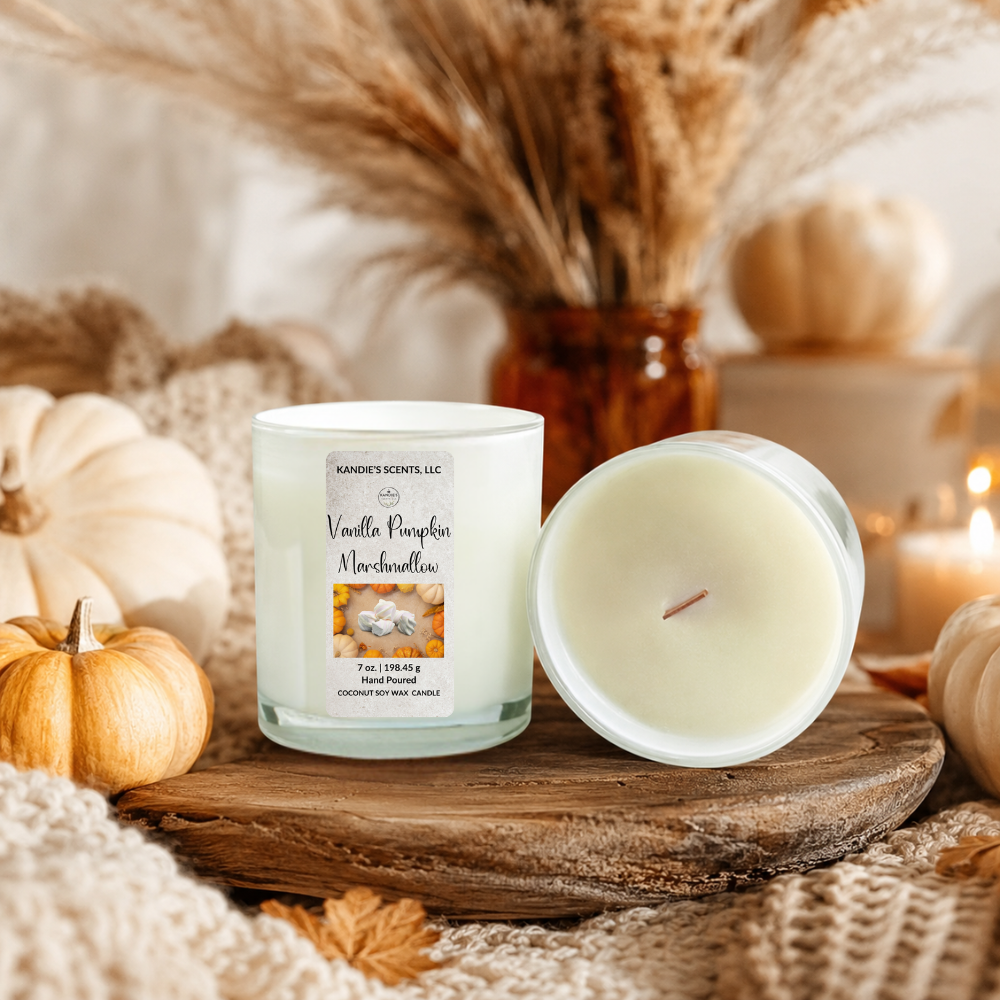 A scented candle in a white glass jar with a white label reading 'Vanilla Pumpkin Marshmallow' by Kandie's Scents, shown next to its open lid on a rustic wooden board, surrounded by pumpkins, dried grass, and a cozy knitted blanket.