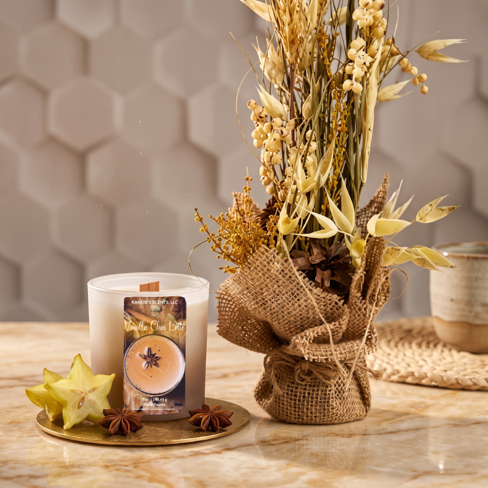 A scented candle in a tan glass jar with a wood wick sits beside a large burlap-wrapped arrangement of dried wheat, millet, and small yellow flowers on a marble surface. Star anise and a yellow starfruit are placed near the candle, with a softly blurred beige hexagonal wall and woven mat in the background.