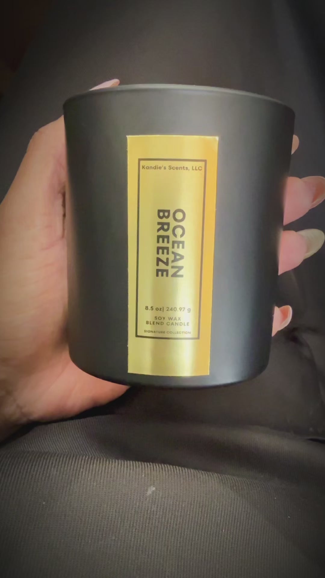 A close-up video of an 8.5 oz Ocean Breeze candle held in one hand and gently rotated to highlight its elegant gold foil label. The candle is housed in a matte black jar, and the reflective label catches the light, showcasing the luxury and craftsmanship of Kandie’s Scents’ signature scent.